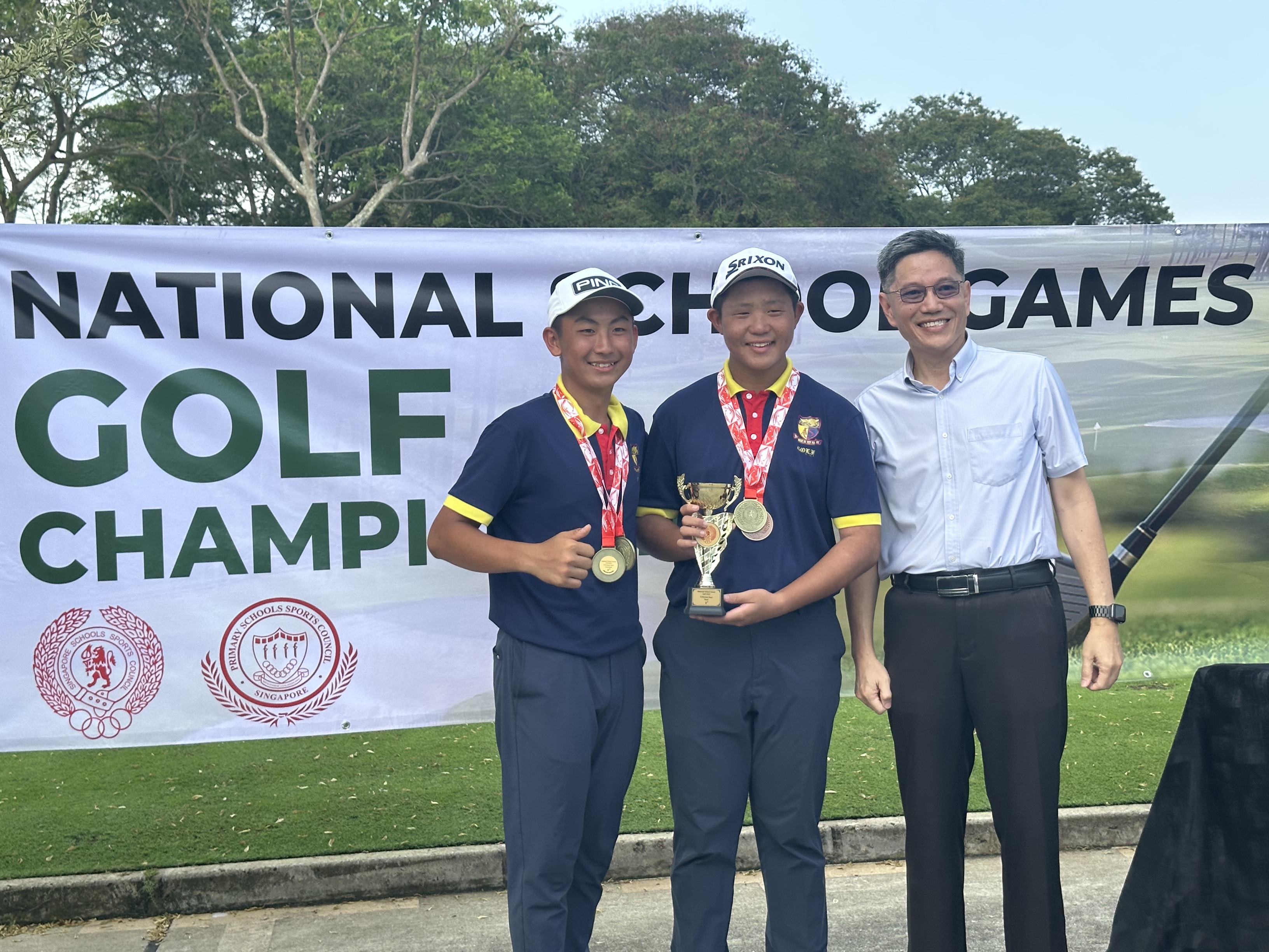 Two young golfers with medals and a trophy pose with an adult before a National School Games Golf Championship banner.